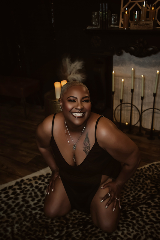 A woman with short blonde hair leans into her knees in front of a candlelit fireplace where she laughs and smiles during her Boudoir session in Hillsboro Missouri.