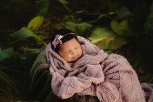 Baby girl covered in mauve colored blanket is nestled into a basket for her outdoor newborn photo session