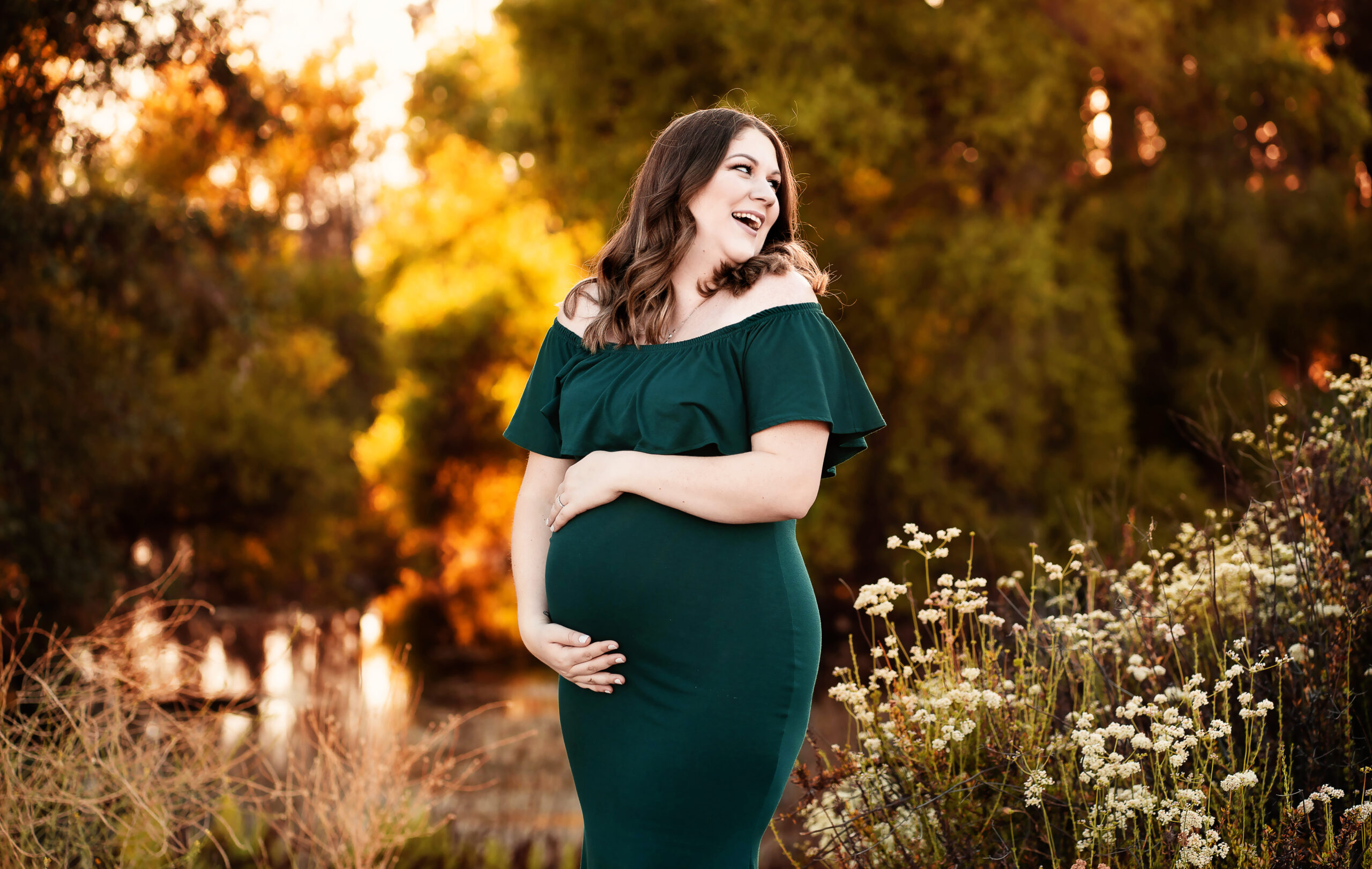 A woman wearing an emerald green maternity dress looks over her shoulder as she laughs and enjoys her maternity photo session with photographer Andrea Moore