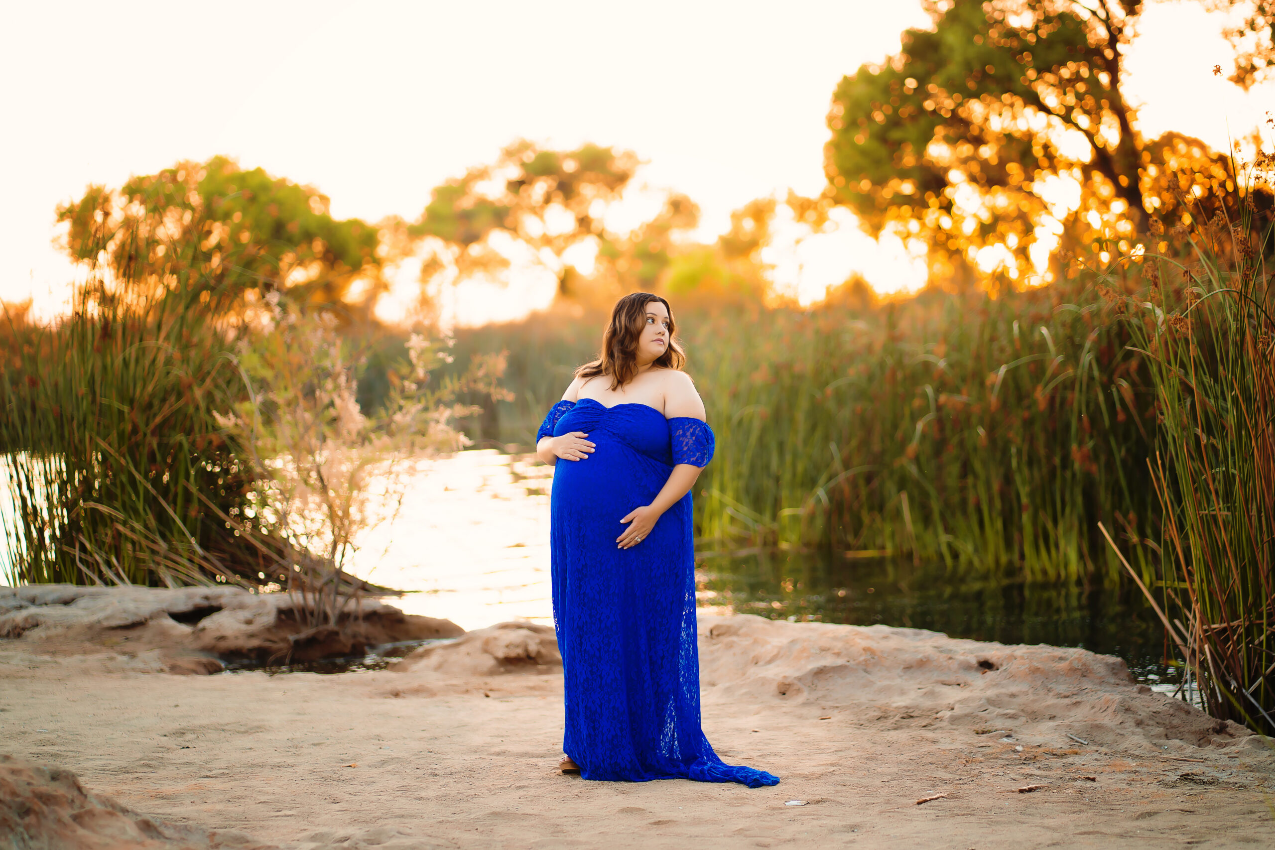 A woman wearing a royal blue maternity gown poses next to a lake as the sunset glows behind her during her maternity photo session