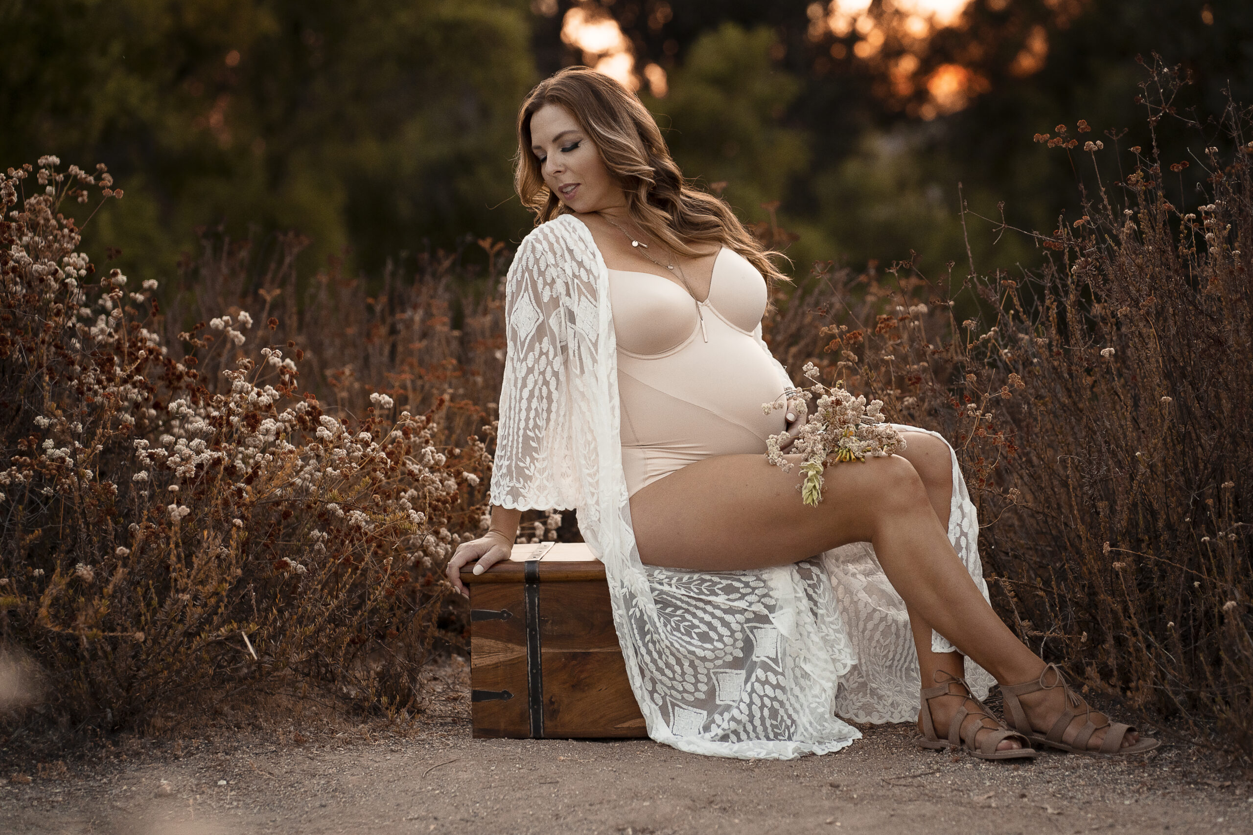 Pregnant woman sits on a wooden trunk in a field full of brown and white flowers while wearing lingerie as she poses for her maternity photo session