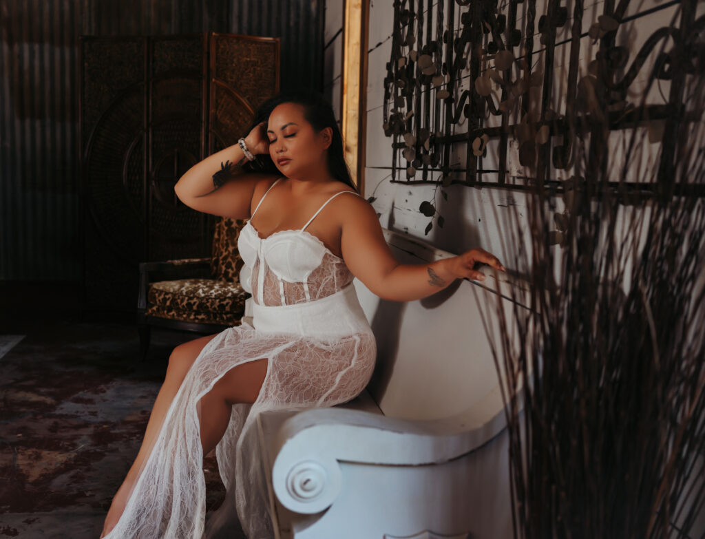 A woman touches her hair as she sits on a white bench during her in studio boudoir photo session.