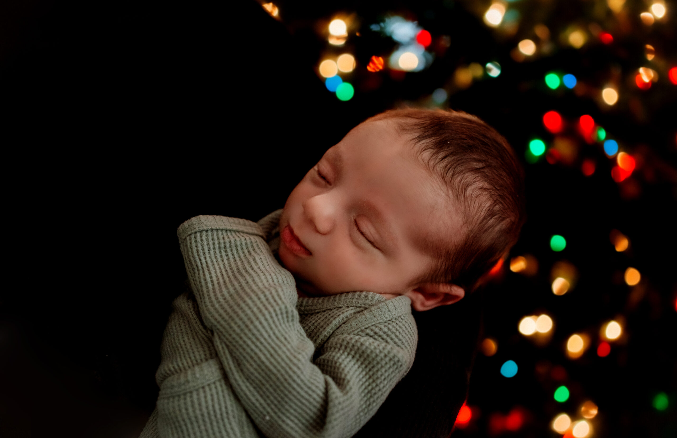 Newborn baby sleeps comfortably in his mother's arms as they pose in front of the glistening lights of the Christmas Tree.