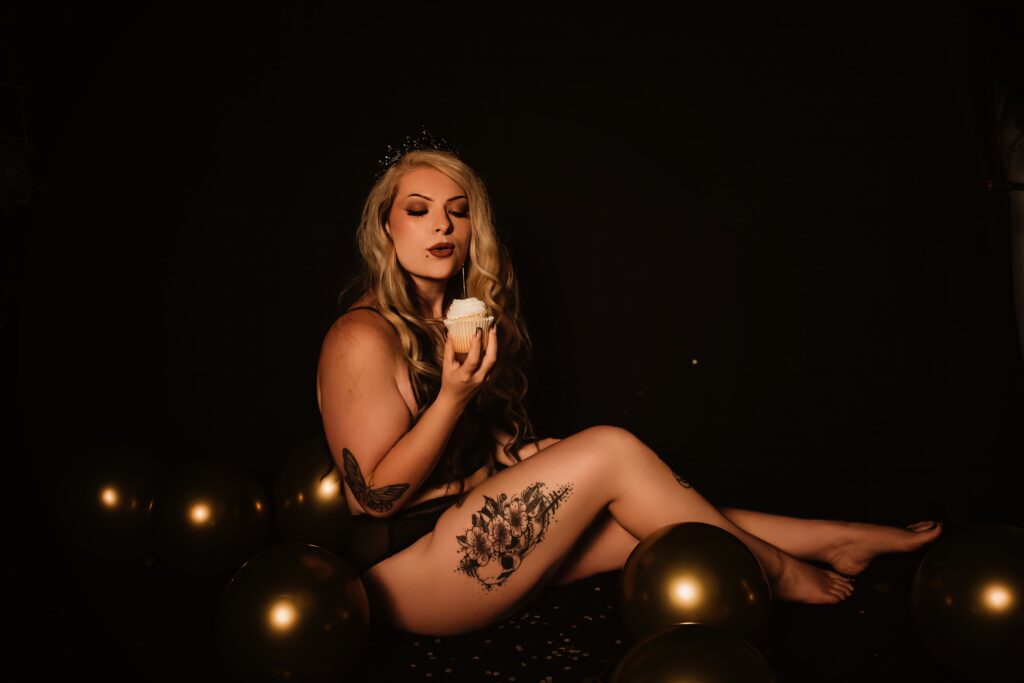 A woman with long blonde hair poses in a photo studio as she blows out the candle on a vanilla cupcake during her boudoir birthday photography session.