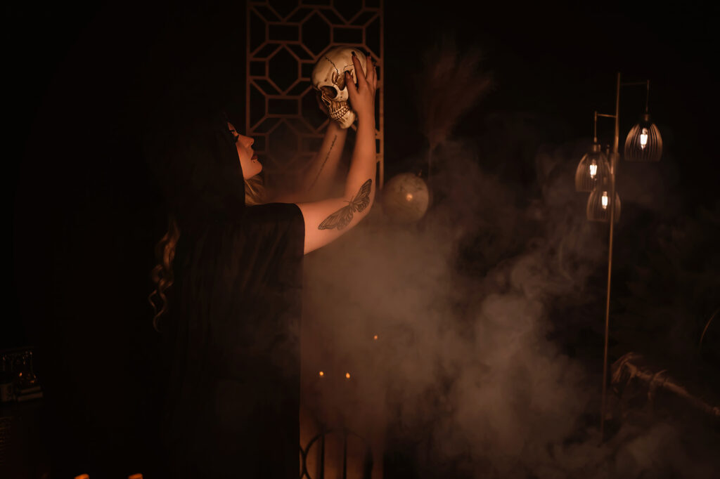 A woman holds a skull above her in a smoke filled room to create an eerie vibe for her Halloween-inspired spooky boudoir session in the St. Louis Missouri area.