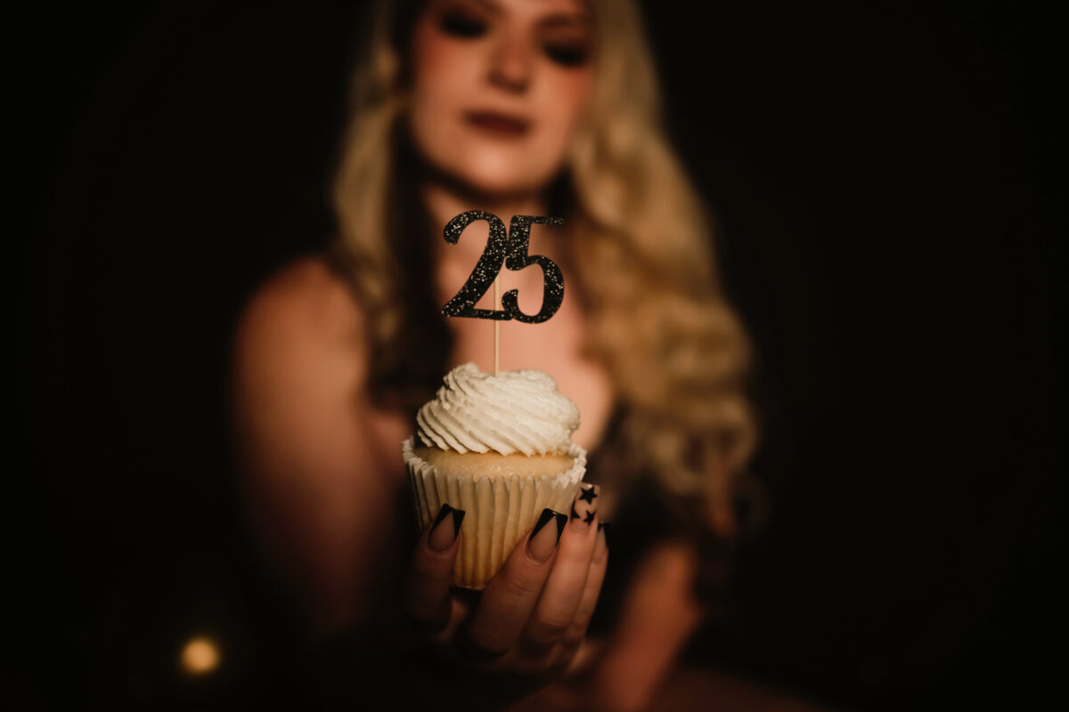 A woman holds a cupcake out in front of her with a decorative "25" on top of it to commemorate her 25th Birthday with an empowering Boudoir Photo Session