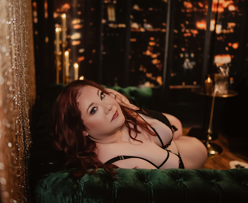 In a beautifully lit image a woman leans on a green velvet sofa as she stares into the camera during her boudoir photography session in Hillsboro Missouri.