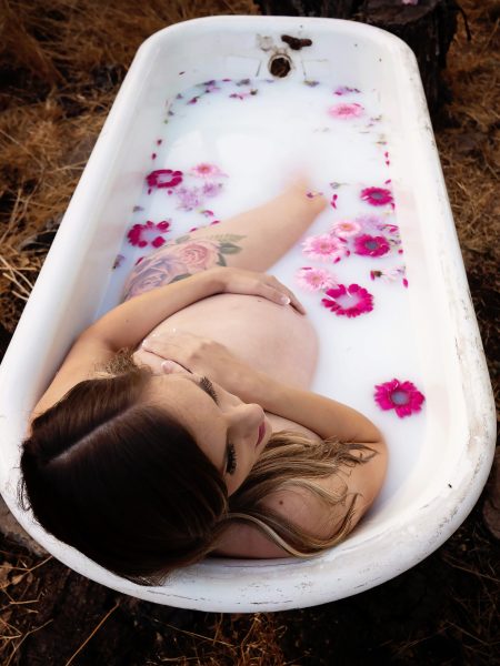 Woman with floral tattoos on her thigh poses in milk bath with pink flowers during her boudoir maternity photo session with photographer Andrea Moore.