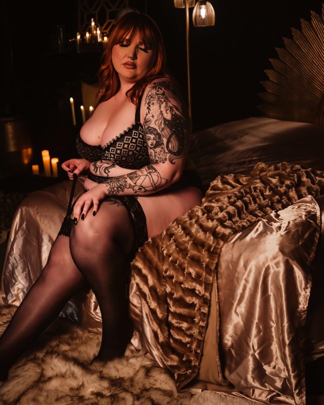 A woman sits on the end of a bed with a gold sun wall decor behind her as she wears black lingerie and knee highs, for her boudoir photo session.