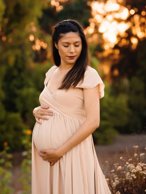 An expecting mother wearing a formal beige maternity gown closes her eyes and holds her pregnant belly during her maternity photo session
