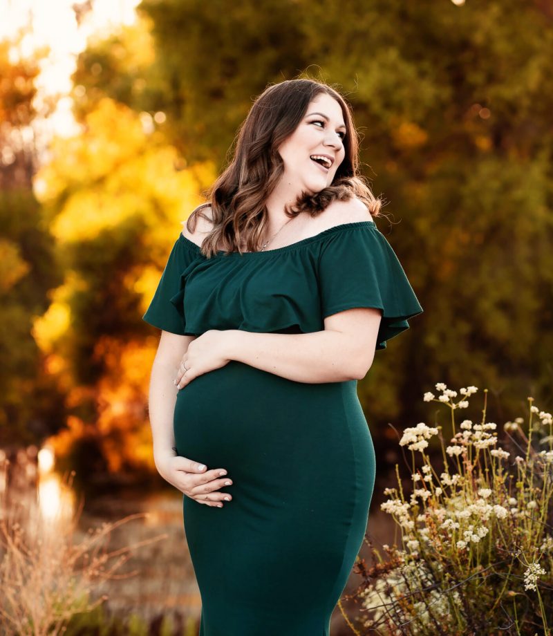 A woman wearing an emerald green maternity dress looks over her shoulder as she laughs and enjoys her maternity photo session with photographer Andrea Moore