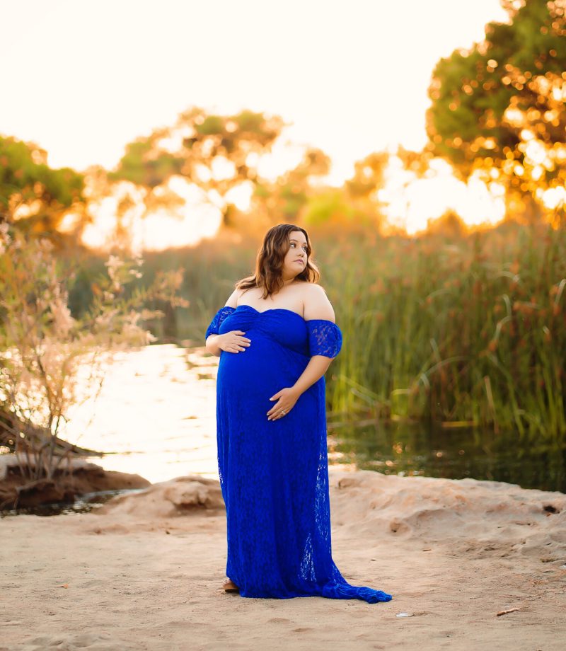 A woman wearing a royal blue maternity gown poses next to a lake as the sunset glows behind her during her maternity photo session