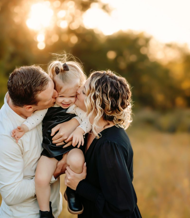 Parents of a young girl give her kisses on the cheeks as they squeeze in close during their sunset family photo session with Love to the Moon Photography near St. Louis Missouri.