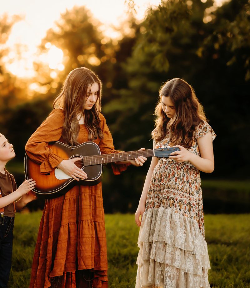 Three siblings hold onto a guitar while standing in front of a pond at sunset during their family photo session with Love to the Moon Photography near Festus Missouri.