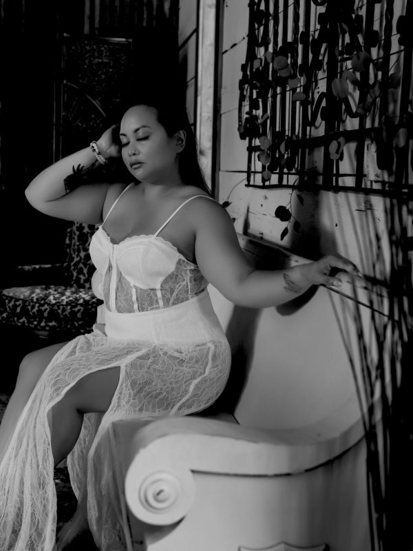 In a black and white photo a woman wearing white lingerie gently closes her eyes as she raises her arm to her head during her boudoir photo session.