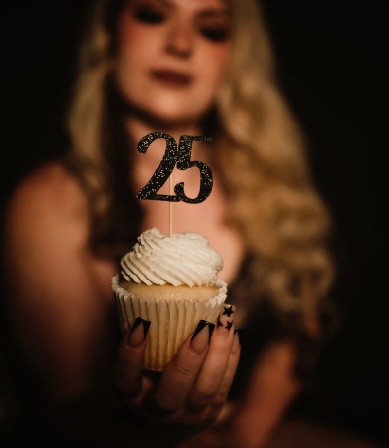 A woman holds a cupcake out in front of her with a decorative "25" on top of it to commemorate her 25th Birthday with an empowering Boudoir Photo Session