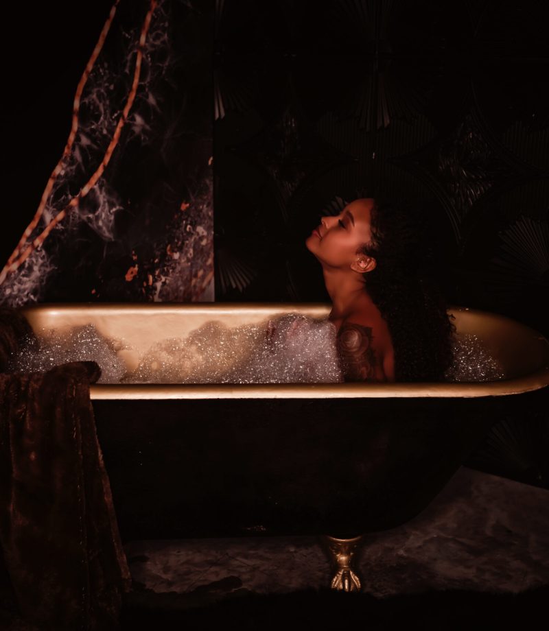 A woman with long dark hair sits in a black and gold claw foot tub full of bubbles, during her boudoir photography session with Love to the Moon Photography near St. Louis Missouri.