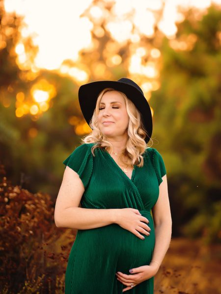 A blonde haired woman in a green dressholds her pregnant belly as she closes her eyes in a field at sunset.