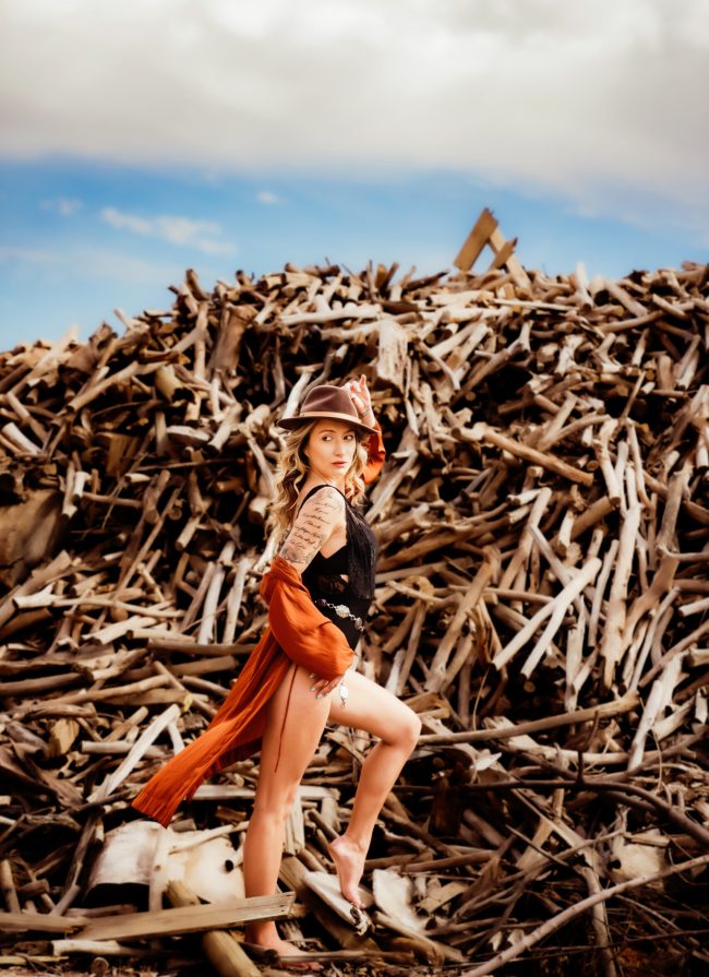A woman wearing a black bodysuit, and an orange coverup with a black hat poses in front of a giant pile of sticks during her boudoir photo session with St Louis based Photographer Love to the Moon Photography