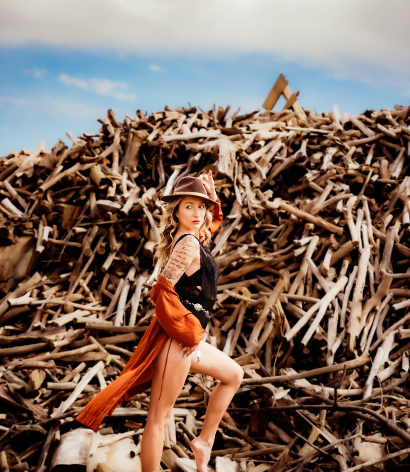 A woman wearing a black bodysuit, and an orange coverup with a black hat poses in front of a giant pile of sticks during her boudoir photo session with St Louis based Photographer Love to the Moon Photography