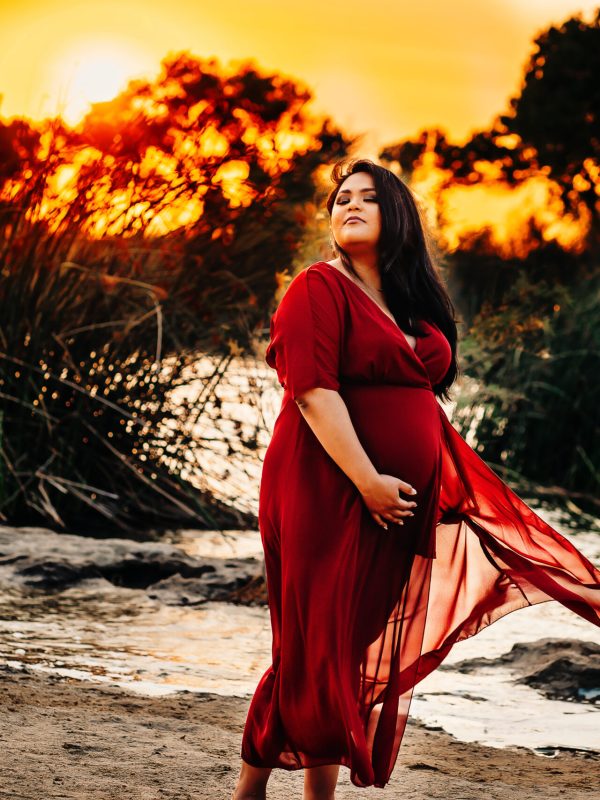 A woman wearing a red dress poses in front of the lake at sunset as her dress flows in the wind during her maternity photo session
